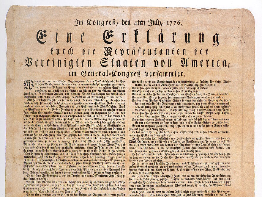 Historisches Druckdokument auf vergilbtem Papier. Überschrift in Frakturschrift: „Im Congreß, den 4ten July, 1776. Eine Erklärung durch die Repräsentanten der Vereinigten Staaten von America, im General-Congreß versammlet.“ Darunter engbedruckter Fließtext in zwei Spalten, am unteren Rand Unterschriften, darunter „John Hancock, Präsident“. Das Papier zeigt deutliche Alterungsspuren mit leichter Bräunung und unregelmäßigen Rändern.