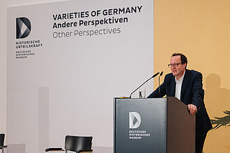 Mann in dunklem Anzug steht am Rednerpult mit Logo des Deutschen Historischen Museums, im Hintergrund Wand mit Text 