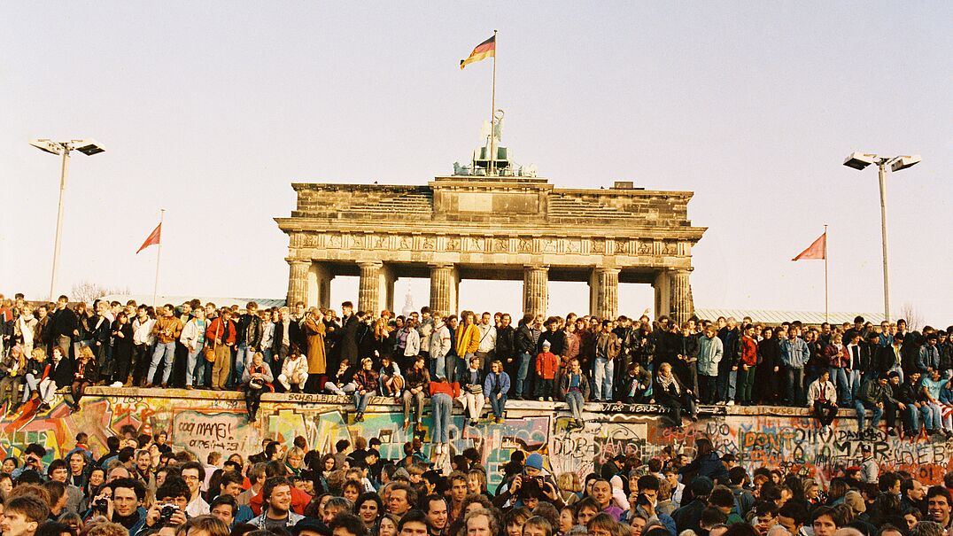 Menschen stehen vor und auf der Berliner Mauer und im Hintergrund ist das Brandenburger Tor.