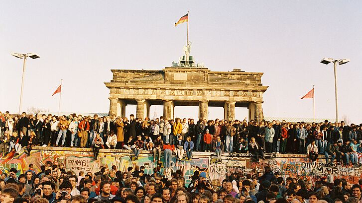 Menschen stehen vor und auf der Berliner Mauer und im Hintergrund ist das Brandenburger Tor.