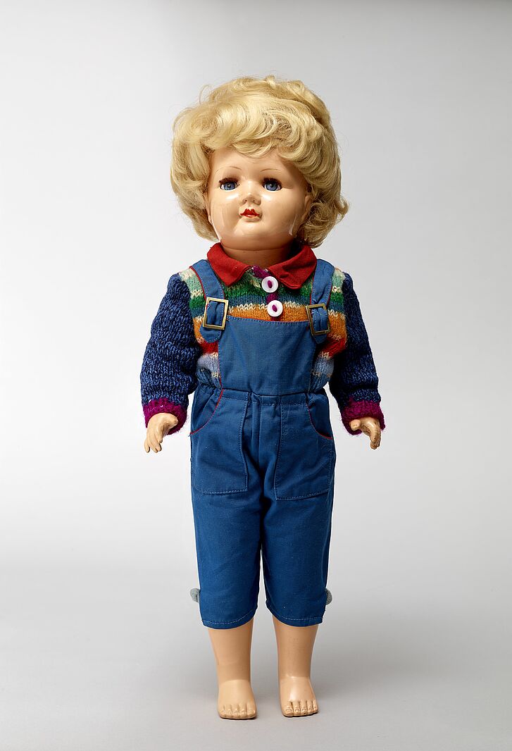 Puppe, mit Latzhose und Pullover bekleidet.