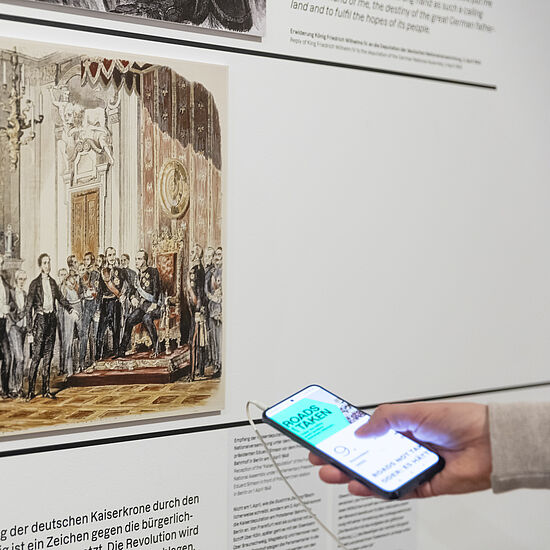 Eine Person hat ein Handy in der Hand, mit der sie den Medienguide bedient und vor einer Wand mit Bild und Text steht.