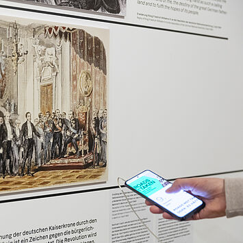 Eine Person hat ein Handy in der Hand, mit der sie den Medienguide bedient und vor einer Wand mit Bild und Text steht.