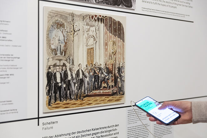 Eine Person hat ein Handy in der Hand, mit der sie den Medienguide bedient und vor einer Wand mit Bild und Text steht.