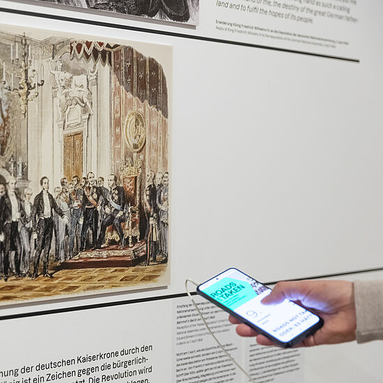 Eine Person hat ein Handy in der Hand, mit der sie den Medienguide bedient und vor einer Wand mit Bild und Text steht.