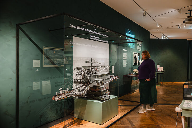 Ausstellungsansicht "Natur und deutsche Geschichte. Glaube – Biologie – Macht" Eine Frau in einem violetten Pullover und grünem Rock betrachtet einen Bagger in einer Vitrine.