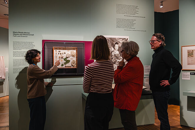 [Translate to English:] Personen stehen in einer Ausstellung und hören einer Referentin zu