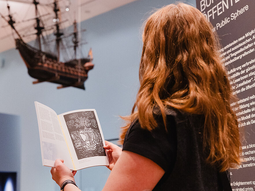 Person, von hinten fotografiert, hält Buch mit Druckgrafik eines Segelschiffs hoch; dahinter hängt ein Schiffsmodell. Rechts ist eine Ausstellungstafel mit der Aufschrift „Bürgerliche Öffentlichkeit“ zu sehen.