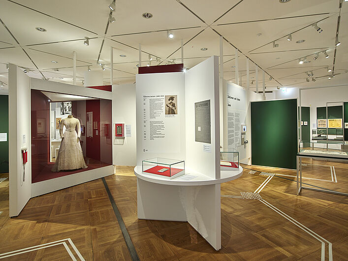 Auf dem Bild ist eine Szene der Ausstellung zu sehen. Es handelt sich dabei um eine Raumansicht. Links im Bild ist ein weißes Hochzeitskleid in einer Vitrine zu sehen. Auf dem Boden das Blindenleitsystem.