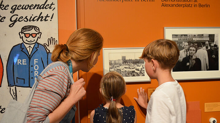 Eine Frau und zwei Kinder betrachten ein Foto, das an der orangenen Ausstellungswand hängt.