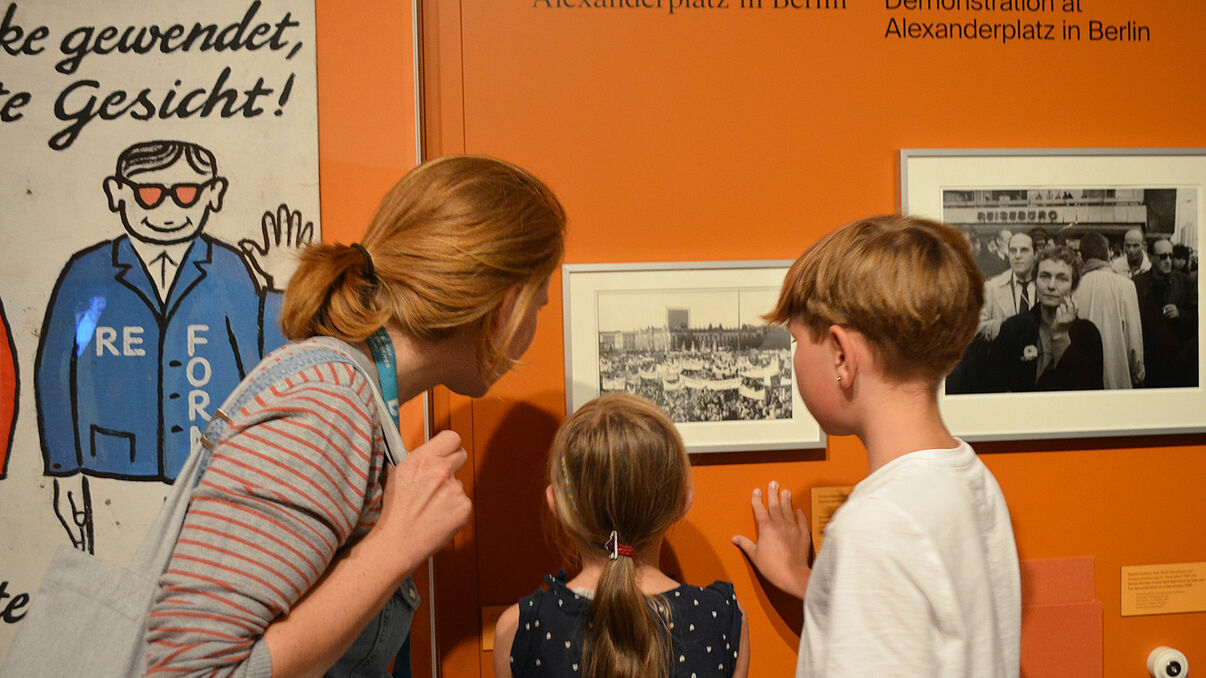 Kinderführung "Warte nicht mit deinem Mut!" Eine Frau und zwei Kinder betrachten ein Foto, das an der orangenen Ausstellungswand hängt.