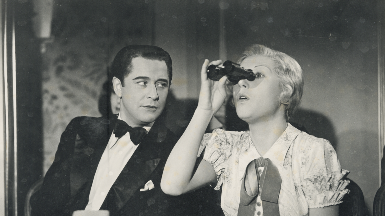 Eine Frau blickt in ein Fernglas und ein Mann sitzt neben ihr und betrachtet sie.