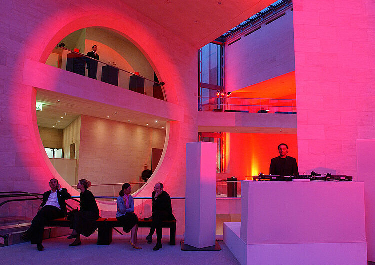 Menschen sitzen und stehen bei abendlicher Beleuchtung im Foyer.