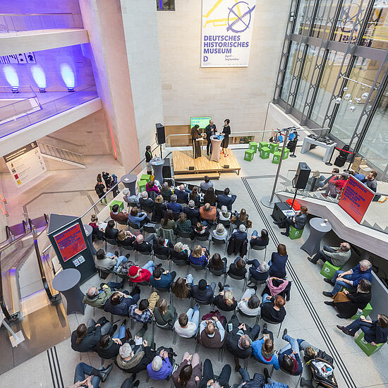 Menschen sitzen vor einer Bühne und hören drei Personen bei einem Gespräch zu.