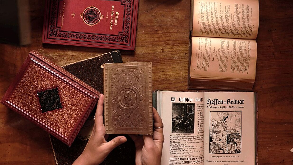 Hände, die ein Buch halten an einem Tisch mit mehreren historischen Büchern.