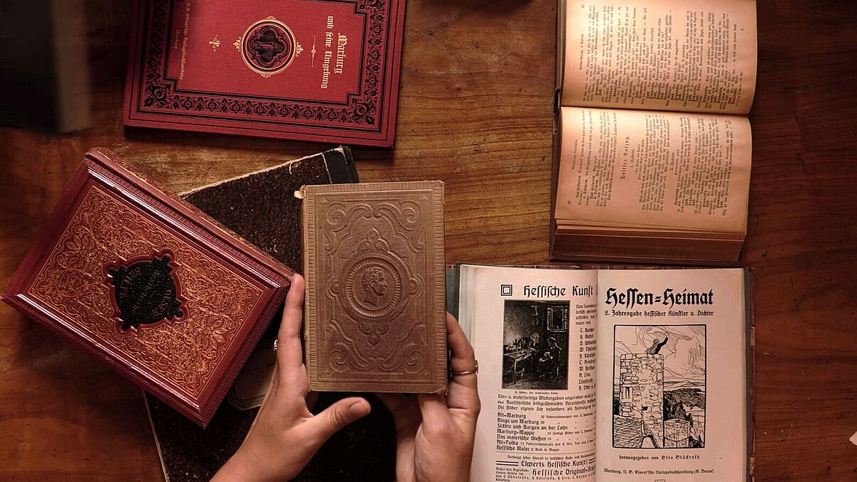 Hände, die ein Buch halten an einem Tisch mit mehreren historischen Büchern.