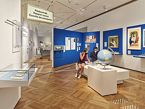 Auf dem Bild sind Personen in der Ausstellung zu sehen. Im Hintergrund sind blaue Wände zu sehen, an denen Objekte hängen. Im Vordergund sitzen Personen und gucken sich Objekte an. Ein Schild mit der Aufschrift "Passkontrolle" ist links im Bild zu sehen.