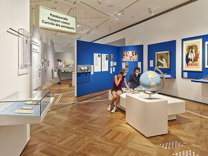 Auf dem Bild sind Personen in der Ausstellung zu sehen. Im Hintergrund sind blaue Wände zu sehen, an denen Objekte hängen. Im Vordergund sitzen Personen und gucken sich Objekte an. Ein Schild mit der Aufschrift "Passkontrolle" ist links im Bild zu sehen.