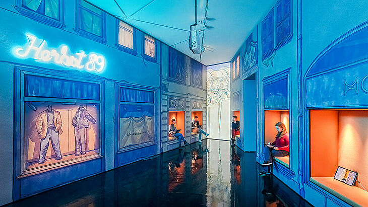 Besucher, die in den Sitzecken sitzen, lesen und sehen sich Wandbilder von Schaufensteransichten vom Herbst 1989 an.