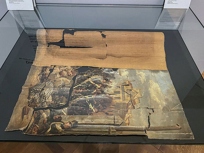 Ein zerrissenes, historisches Gemälde, welches aus dem Rahmen genommen wurde und auf einem Tisch liegt.