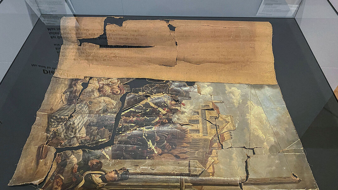 Ein zerrissenes, historisches Gemälde, welches aus dem Rahmen genommen wurde und auf einem Tisch liegt.