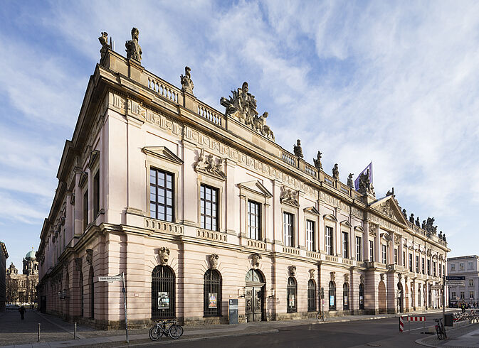 Zeughaus Auf dem Foto ist das Zeughaus zu sehen. Es handelt sich dabei um ein großes Gebäude, das auf dem Bild hellrosa aussieht. Es ist also sehr hell. Auf dem Gebäude sind Figuren zu sehen.