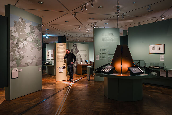 Exhibition view "Nature and German History. Faith – Biology – Power" A man in a black sweater holds the earpiece of a media station to his ear.