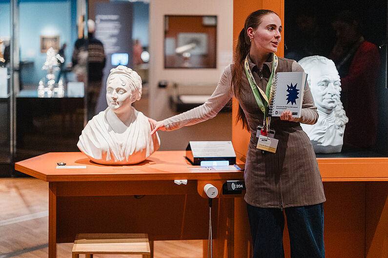 Eine Frau steht neben einer Büste einer anderen Frau in einer Ausstellung und zeigt auf diese.