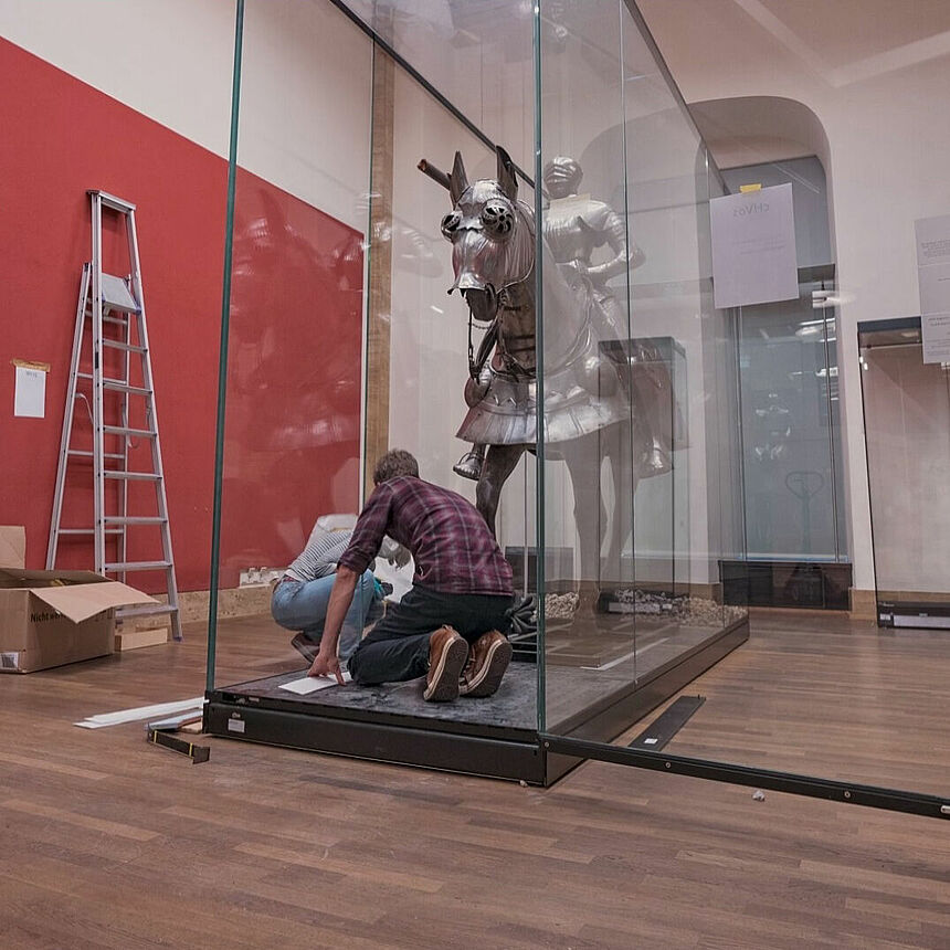 Zwei Personen installieren eine lebensgroße Reiterskulptur in einer Vitrine.