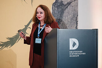 Frau mit rotem Anzug und Namensschild hält eine Brille und steht hinter einem Rednerpult mit dem Logo des Deutschen Historischen Museums.