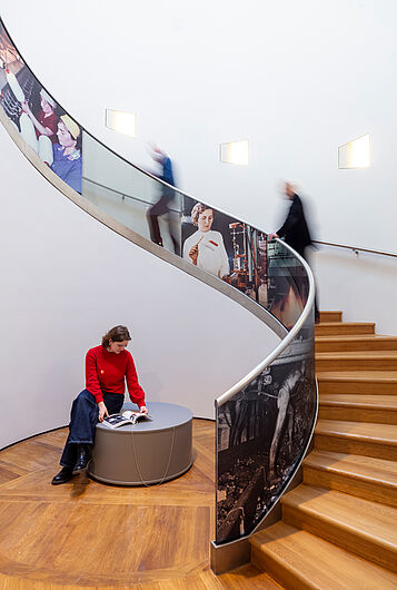 Eine Frau sitzt und blättert in einem Buch und zwei Menschen gehen im Hintergrund eine Treppe hinauf.
