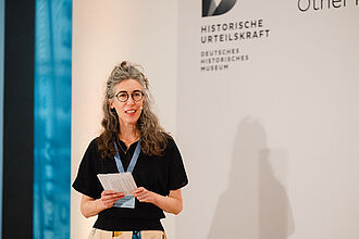 Frau mit langen, gewellten Haaren, schwarzem Oberteil und hellen Hosen hält Zettel vor Wand mit Schriftzug 