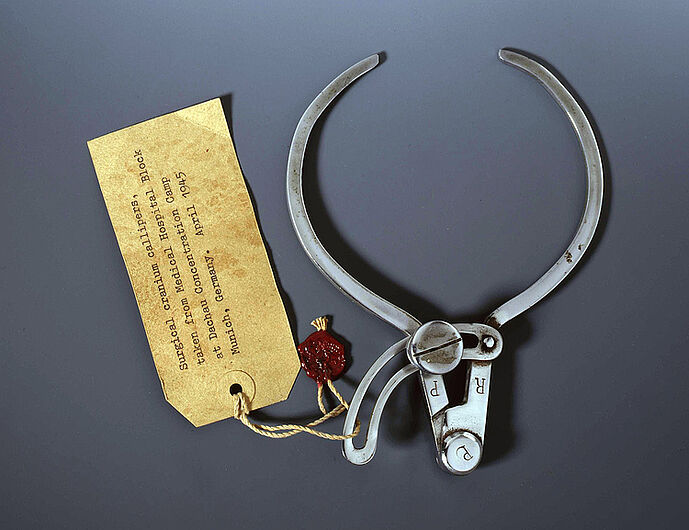 Kraniometer (Instrument zur Vermessung des Schädelumfangs), gefunden im Konzentrationslager Dachau (mit Inventarzettel und Siegel der US-Armee), 1933-1945. Kraniometer (Instrument zur Vermessung des Schädelumfangs), gefunden im Konzentrationslager Dachau (mit Inventarzettel und Siegel der US-Armee), 1933-1945. Kraniometer wurden von Anthropologen seit dem 19. Jahrhundert zur Vermessung von Schädeln genutzt. Im „Dritten Reich“ sollte das Vermessen Aufschluss über die „Rassezugehörigkeit“ von Menschen geben. (Inv.Nr. AK 2001/318)