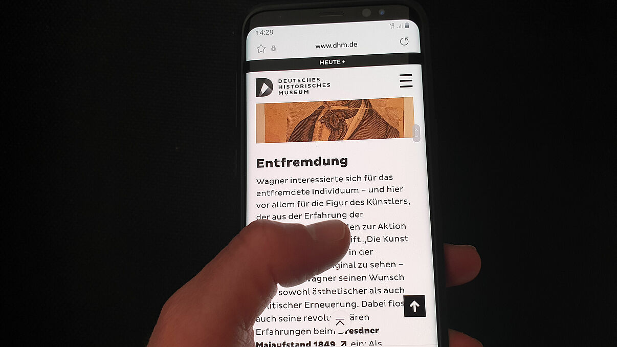 Eine Hand hält ein Smartphone, dass die Website des Formats More Story zur Ausstellung Richard Wagner und das deutsche Gefühl zeigt