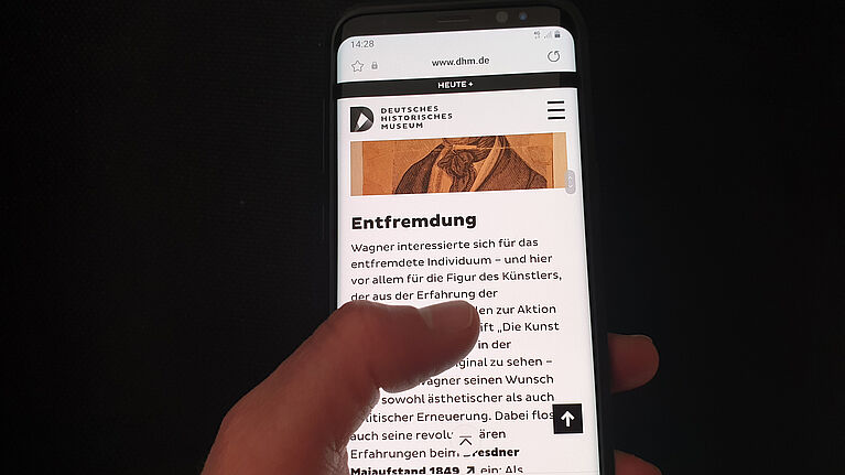 Eine Hand hält ein Smartphone, dass die Website des Formats More Story zur Ausstellung Richard Wagner und das deutsche Gefühl zeigt