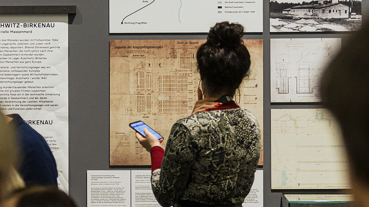 Eine junge Frau steht in einer Ausstellung und hält ein Smartphone in der Hand