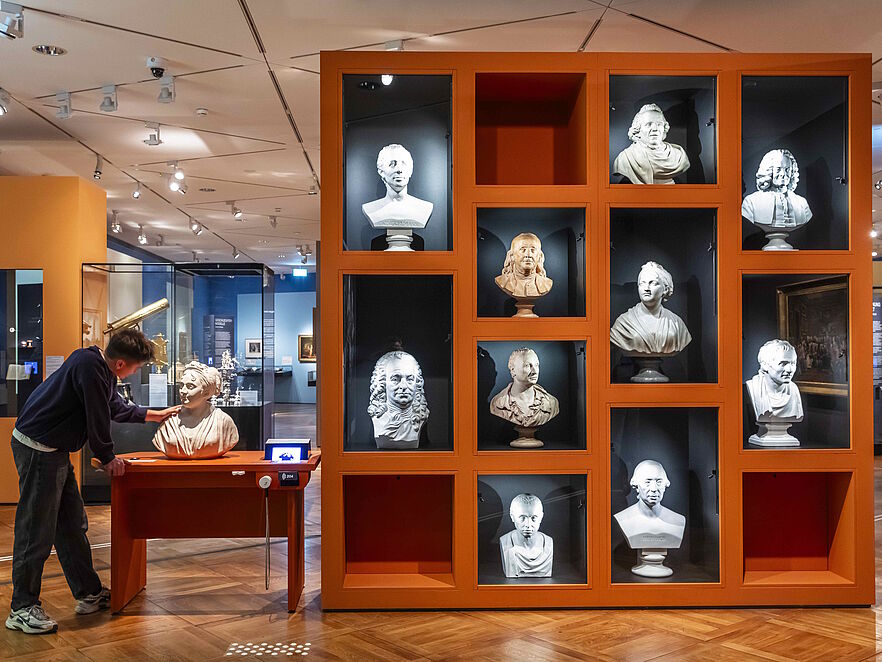 Museumsraum mit orangefarbenem Regal, das zwölf beleuchtete Nischen mit weißen Porträtbüsten zeigt. Ein junger Mann betrachtet links daneben eine einzelne Büste.