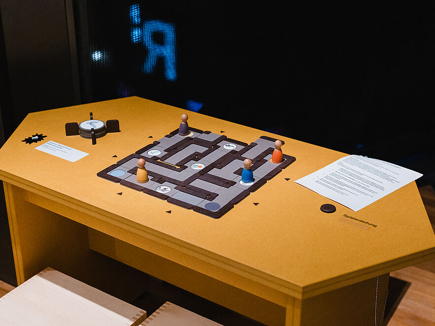 Gelber sechseckiger Tisch in einem abgedunkelten Museumsraum. Auf der Tischfläche liegt ein quadratisches Labyrinth-Brettspiel aus grauem Holz mit dunklen Trennwegen. Vier Spielfiguren in den Farben Beige, Lila, Orange und Blau sind an verschiedenen Positionen auf dem Brettspiel platziert. Rechts liegt ein beschriftetes Blatt über einem Audioknopf mit der Aufschrift „Spielanleitung“.