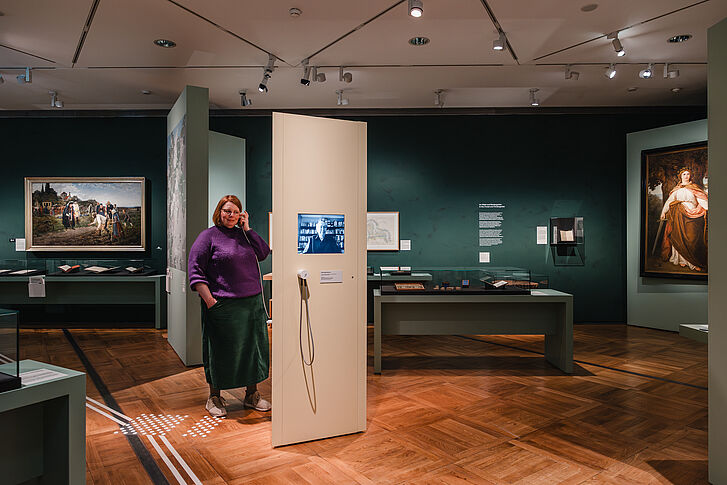 Exhibition view "Nature and German History. Faith – Biology – Power" A woman wearing a purple sweater and a green skirt holds an earpiece from a media station to her ear.