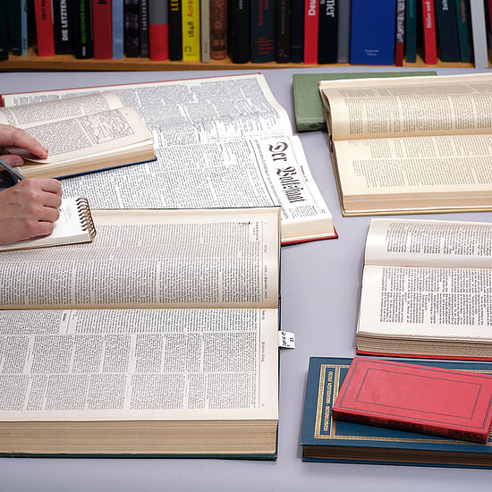 Viele Bücher liegen auf einem Tisch und eine Person macht sich dazu Notizen