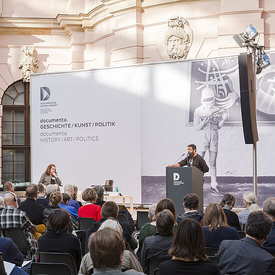 Menschen sitzen vor einer Bühne und hören einem Redner zu.