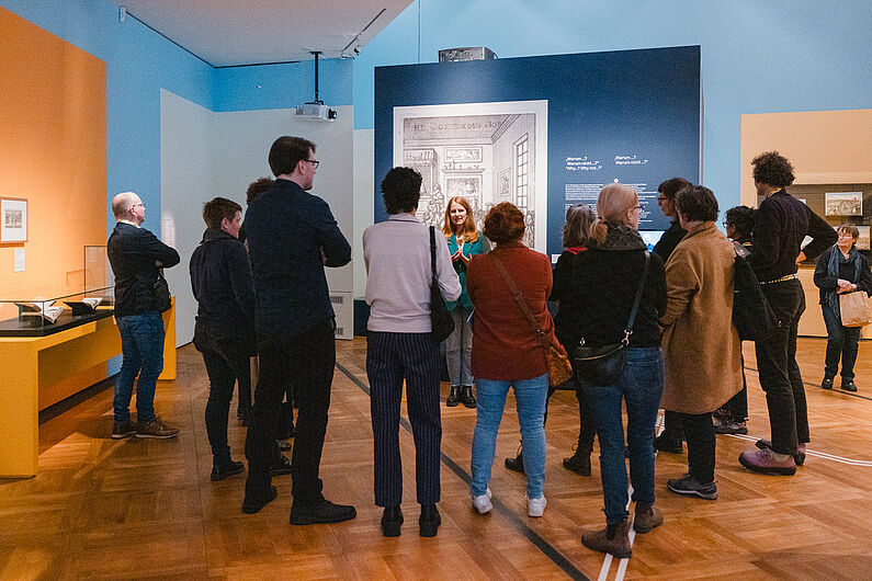 Eine Gruppe von Personen steht in einer Ausstellung und hört einer Frau zu.