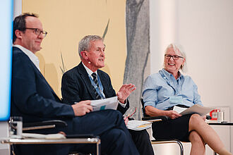 Drei Personen sitzen in einem Gespräch, zwei Männer und eine Frau, alle halten Papiere, im Hintergrund eine Wand mit dem Motiv des Symposiums.