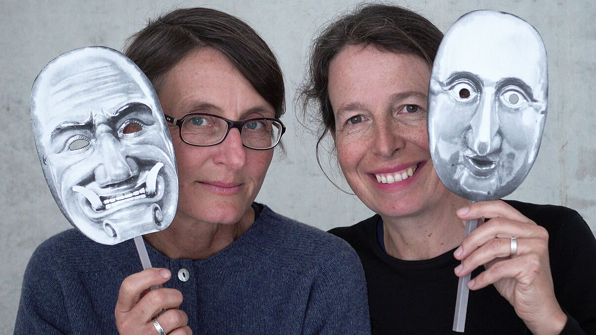 Zwei Frauen mit Masken in der Hand lächeln in die Kamera.