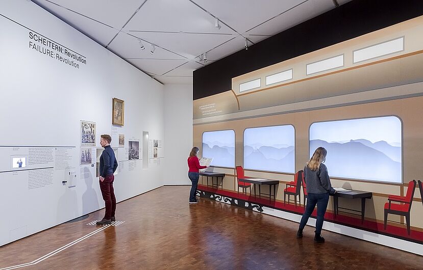 Besucher betrachten eine Museumsausstellung mit einem Wandbild von einem historischen Zug.