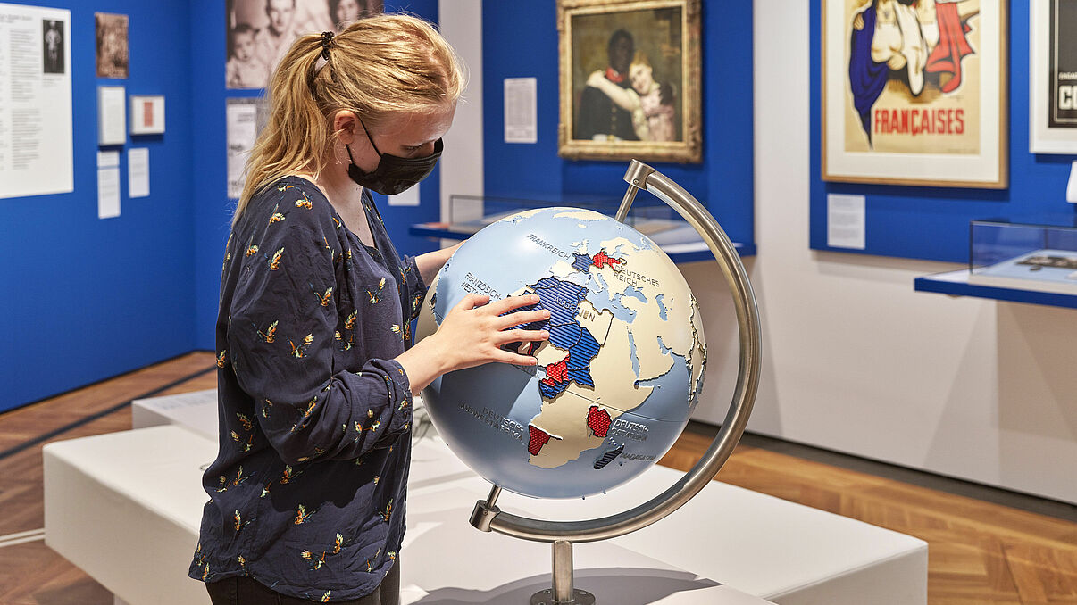 Auf dem Foto ist eine Person in der Ausstellung zu sehen. Die Person guckt auf einen Globus. Der Globus ist ein Glok´bus für Sehbeeinträchtigte Menschen. Einige Länder sind schraffiert, damit man sie erfühlen kann. Im Hintergrund ist ein Gemälde von einem schwarzen Mann und einer weißen Frau zu sehen, die sich im Arm halten.