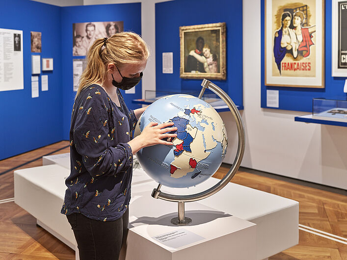 Auf dem Foto ist eine Person in der Ausstellung zu sehen. Die Person guckt auf einen Globus. Der Globus ist ein Glok´bus für Sehbeeinträchtigte Menschen. Einige Länder sind schraffiert, damit man sie erfühlen kann. Im Hintergrund ist ein Gemälde von einem schwarzen Mann und einer weißen Frau zu sehen, die sich im Arm halten.