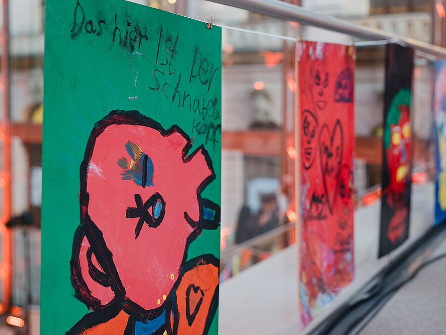 Grünes Plakat mit pinkfarbener Comic-Figur und Aufschrift „Das hier ist der Schnabelkopf“, hängt neben weiteren bunten Kinderzeichnungen an einer Stange in einer Ausstellungshalle.