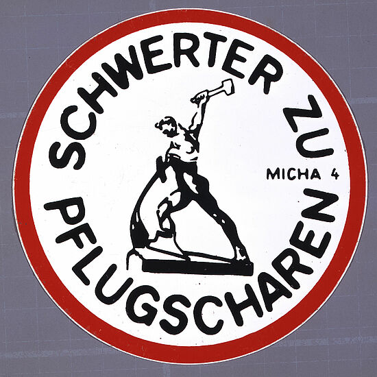 Rundes Abzeichen mit der Aufschrift "Schwerter zu Pflugscharen" und einer Illustration eines Mannes, der ein Schwert zu einem Pflug umschmiedet. Darunter steht "Micha 4", ein Hinweis auf eine Bibelstelle.