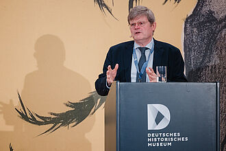 Mann in Anzug und Krawatte spricht hinter einem Rednerpult mit Logo des Deutschen Historischen Museums, im Hintergrund Schatten und Wandzeichnung